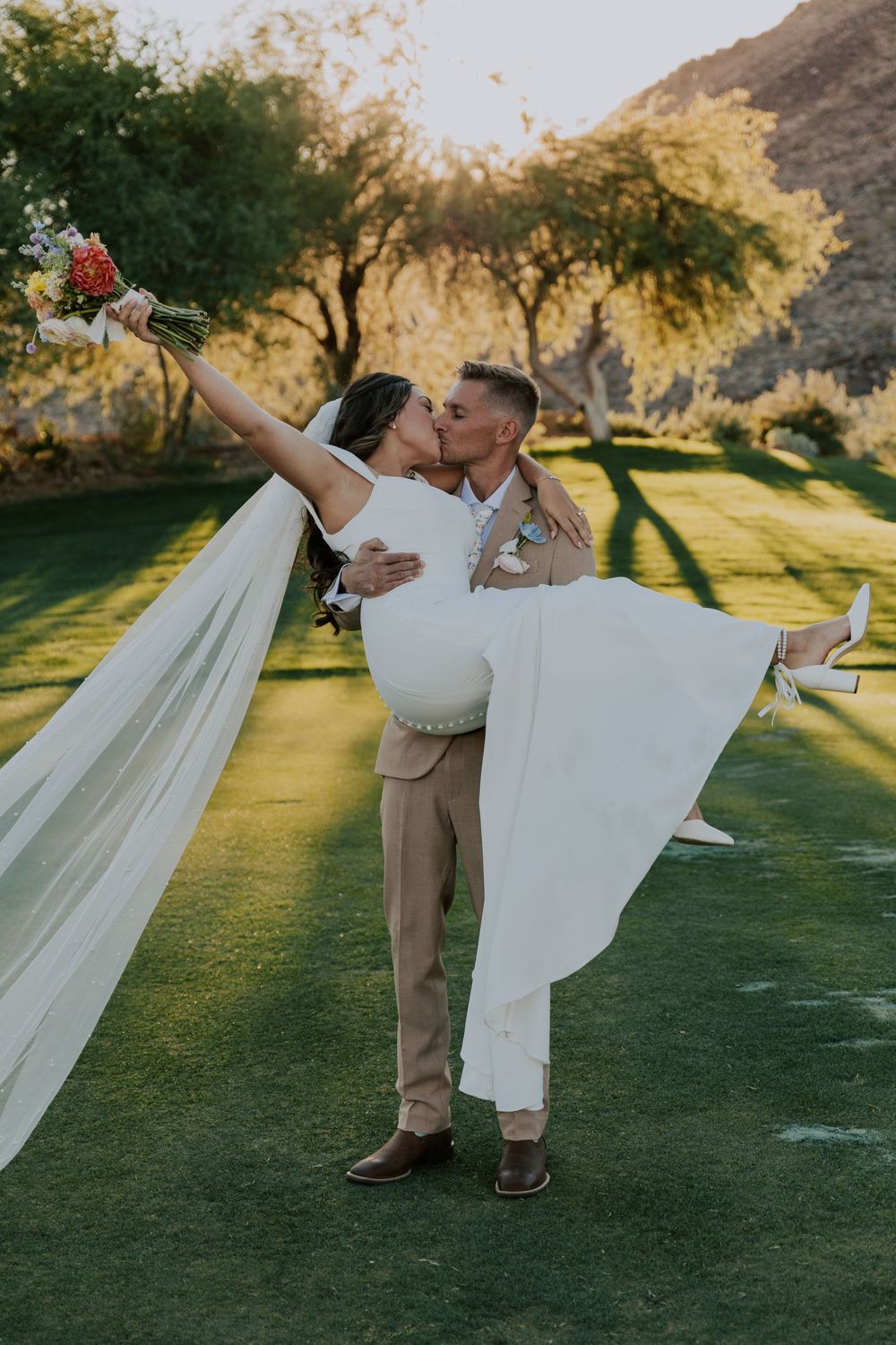 Groom lifts bride in white off-shoulder gown as they kiss on lush green lawn at sunset.