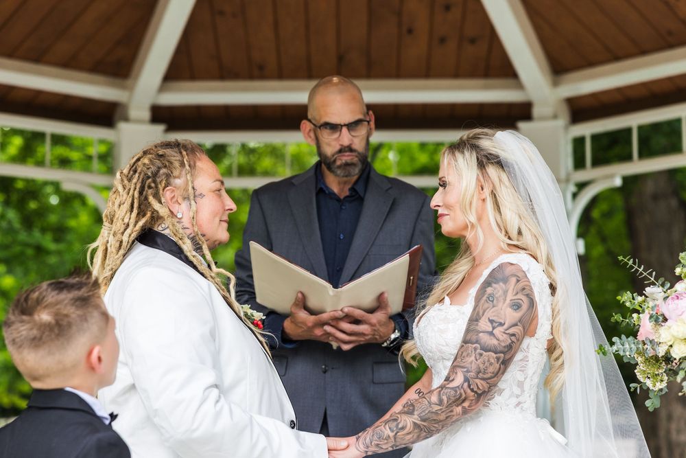 Tattooed bride and dreadlocked bride exchanging vows at outdoor gazebo with officiant.