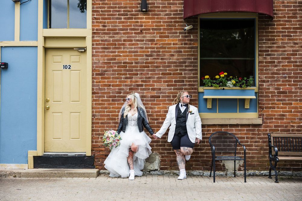 Bride in white gown with leather jacket and bride in white vest walk hand-in-hand past brick building.