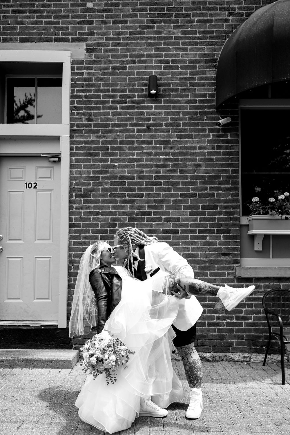 Black and white photo of tattooed bride lifting bride in wedding dress outside brick building.
