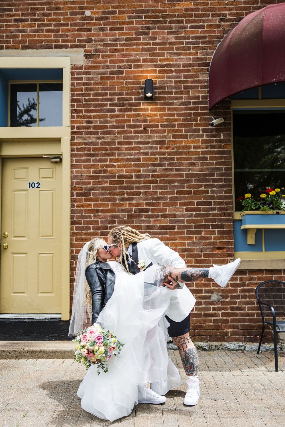 Tattooed bride with dreadlocks dips and kisses bride in white gown outside brick building number 102.