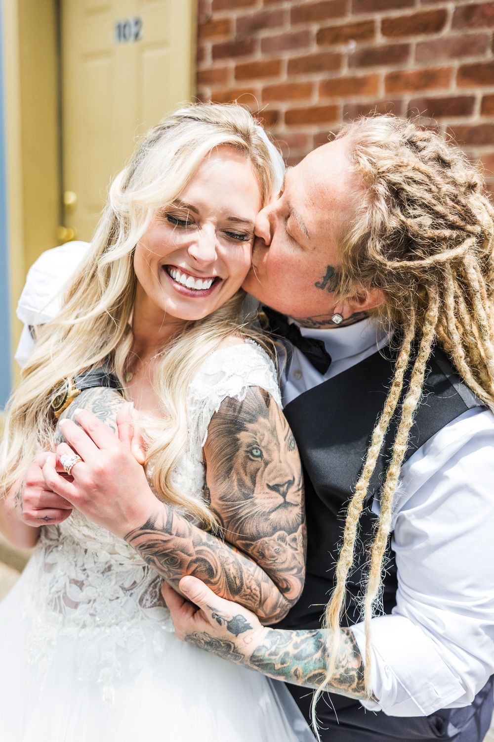 Tattooed bride with dreadlocks kisses smiling blonde bride on the cheek outside brick building.