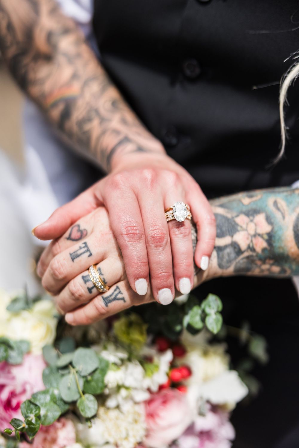 Close-up of bride and bride holding hands showing wedding rings over a pink and white bouquet.