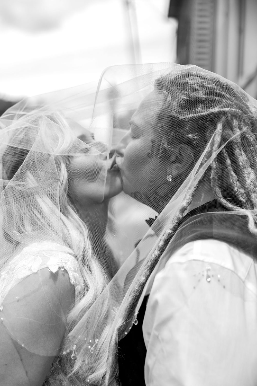 Bride and bride sharing romantic kiss under wedding veil in black and white photo.