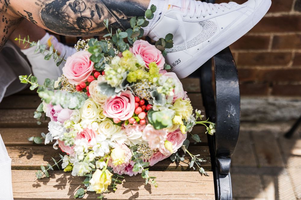 Tattooed bride holding colorful pink rose wedding bouquet with white Nike sneakers on bench.