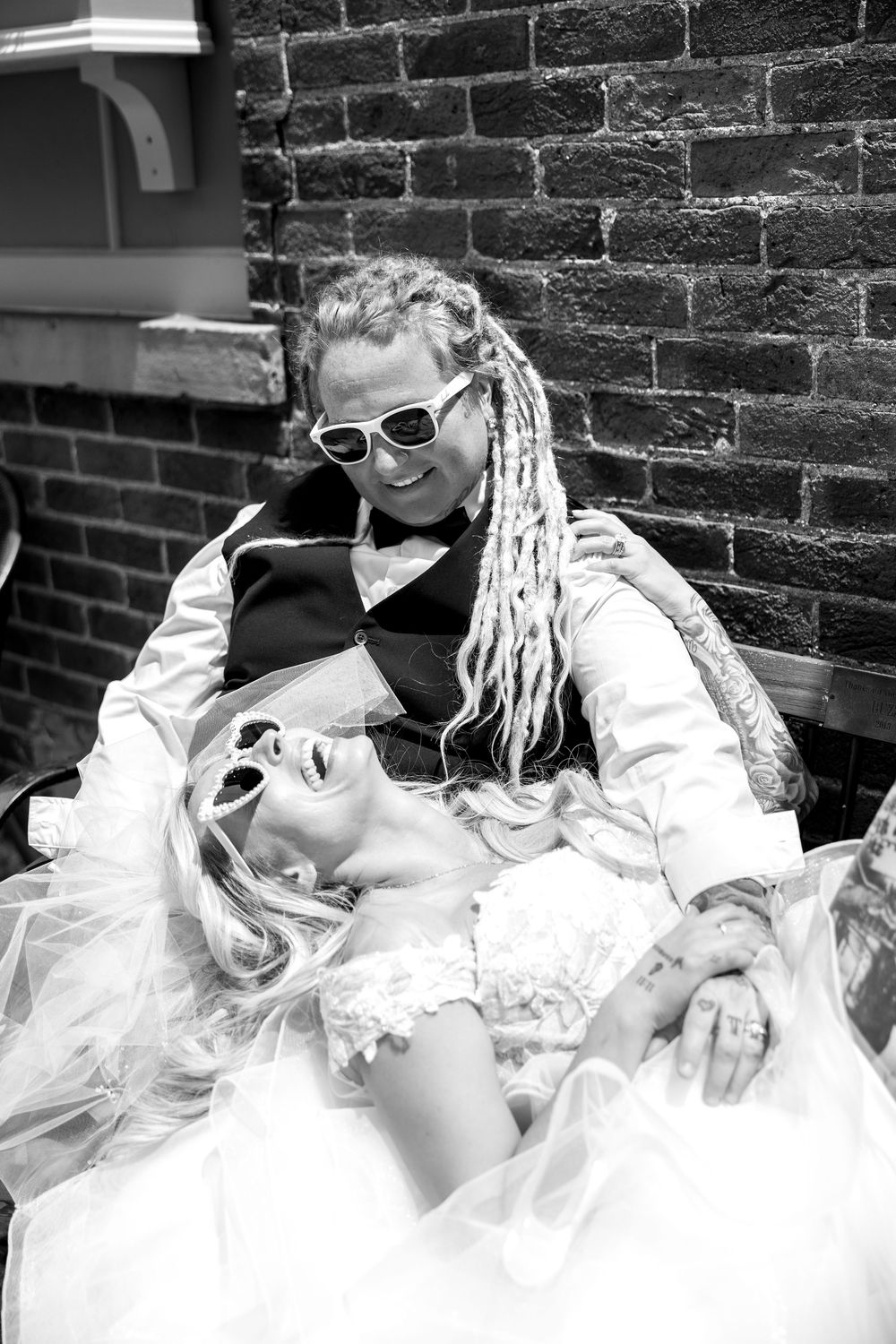 Laughing newlywed couple with dreadlocks and sunglasses posing against brick wall in black and white.