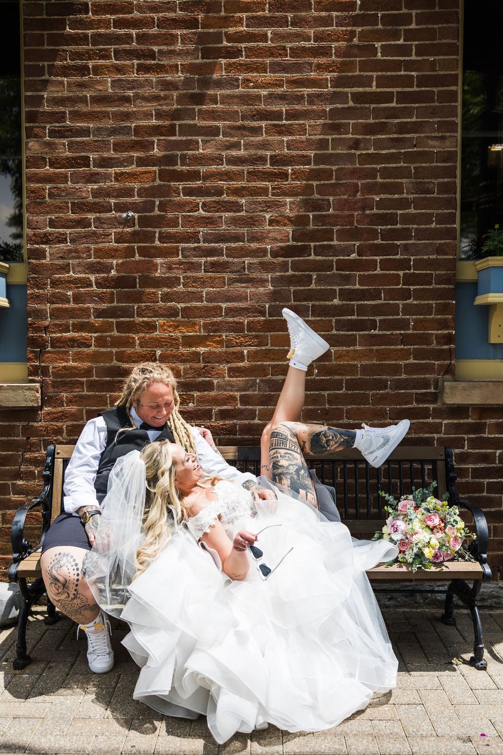 Bride and bride relaxing on bench against brick wall, bride kicking legs up in wedding dress.