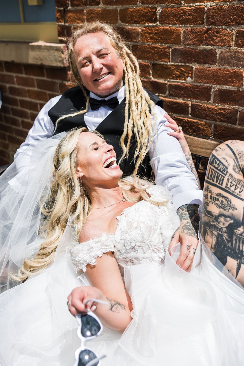 Joyful bride in lace dress laughing with dreadlocked bride holding sunglasses at wedding.