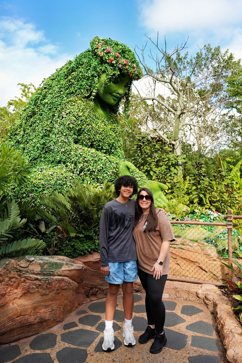 Two visitors pose in front of a giant topiary sculpture of a green leafy figure at Disney's Animal Kingdom theme park.