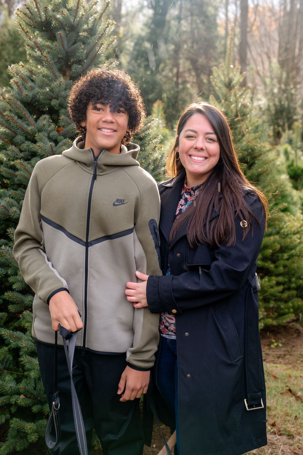 Two people in winter coats standing together at a Christmas tree farm surrounded by evergreen trees.