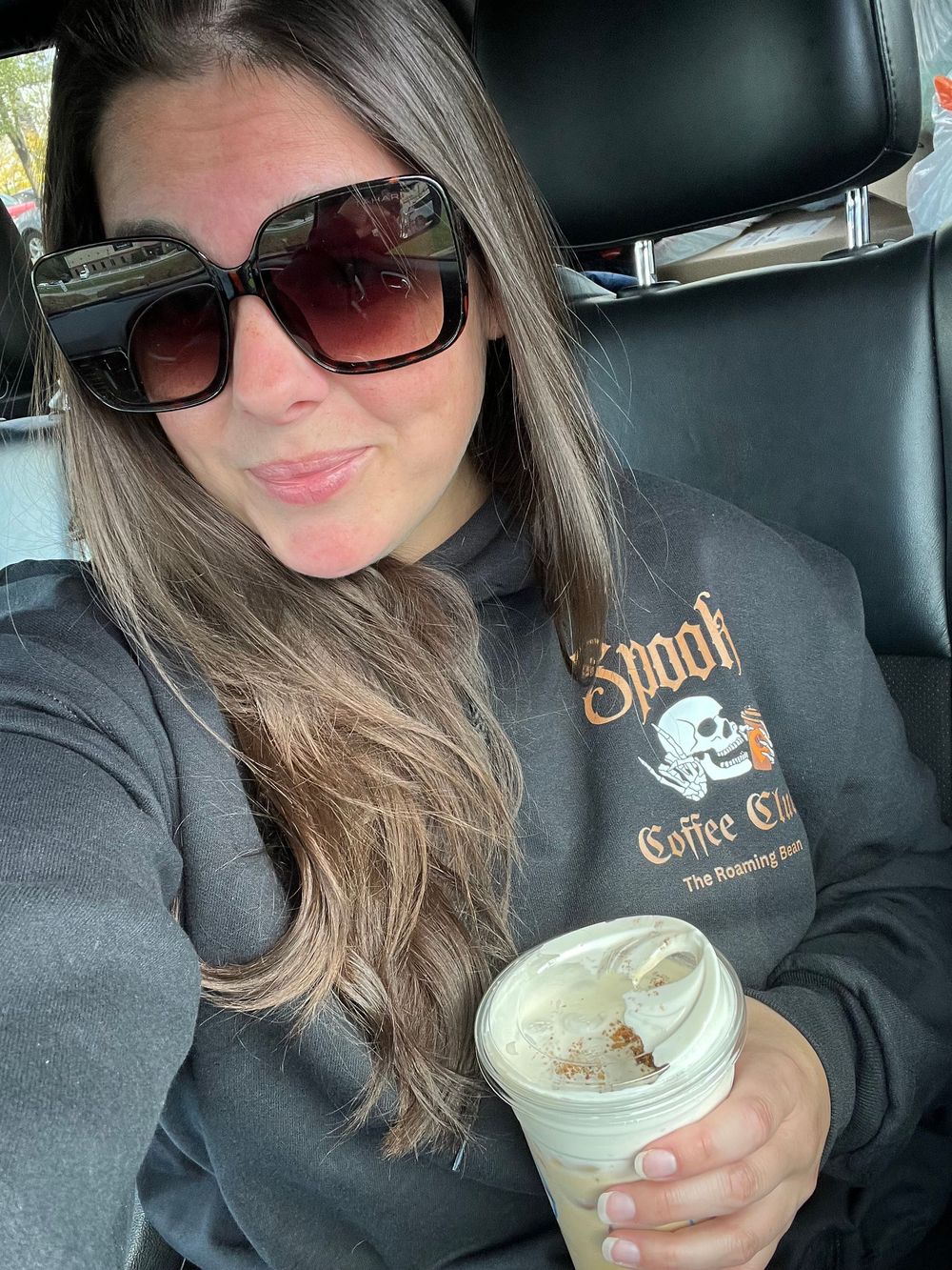 Person enjoying a creamy coffee drink in a car while wearing sunglasses and a black sweatshirt.