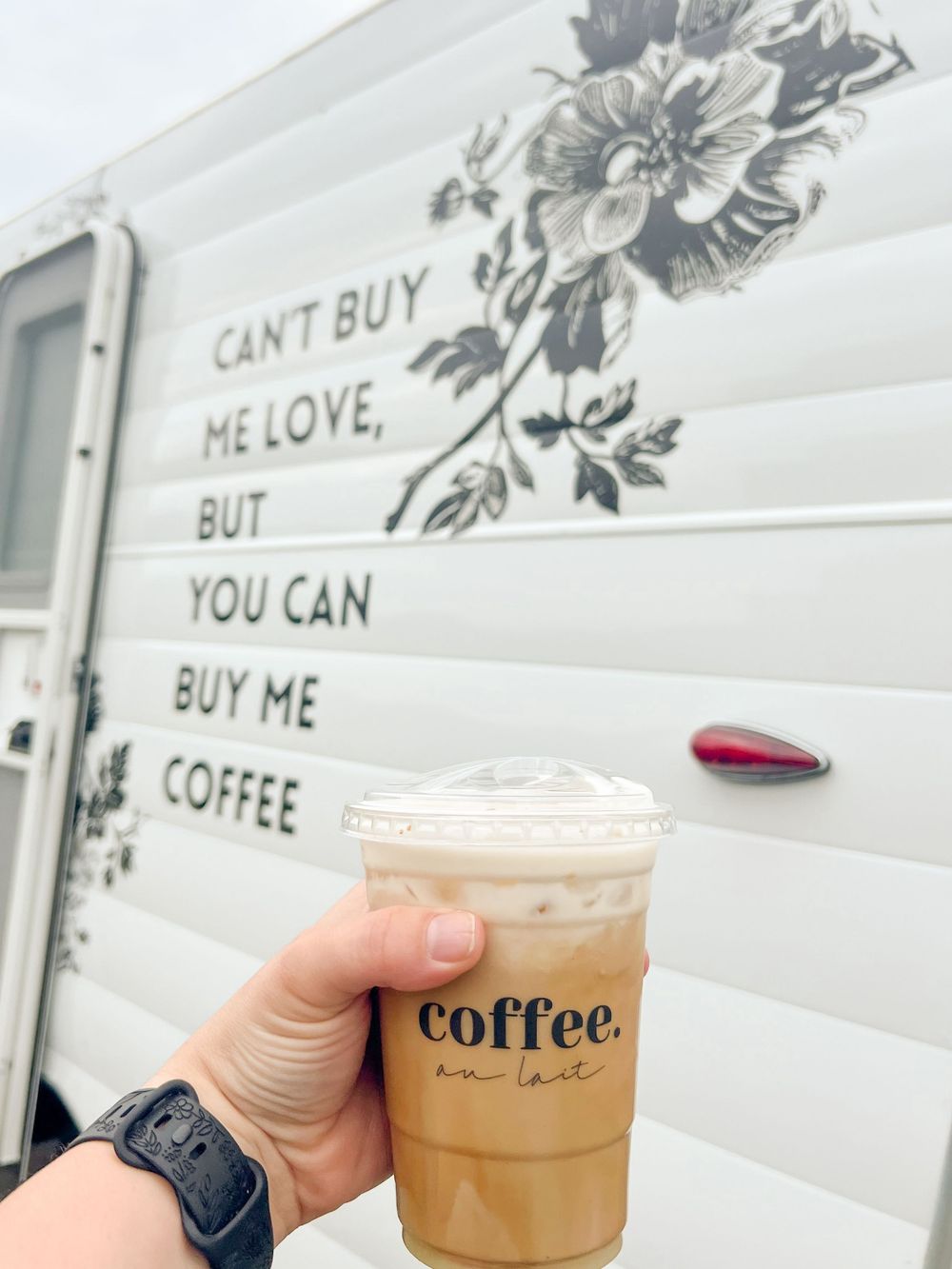 A series of iced coffee drinks in clear plastic cups being held up against a white floral wall background.