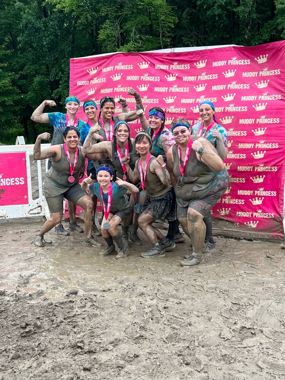 A group of participants poses together in muddy conditions at an outdoor fitness event with pink branded banners.
