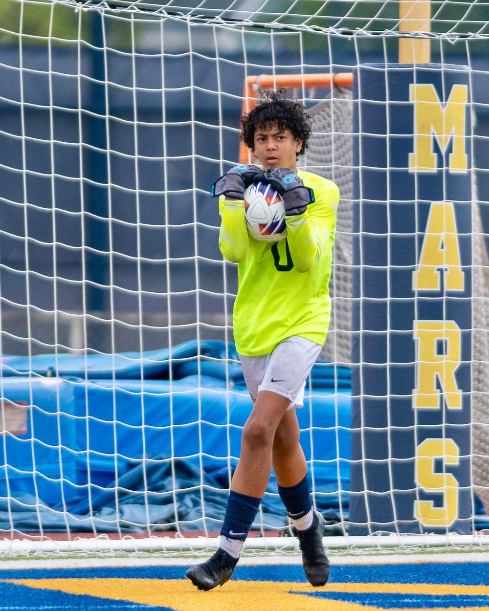 Soccer goalkeeper in bright yellow jersey catches ball while positioned in front of blue net during match.