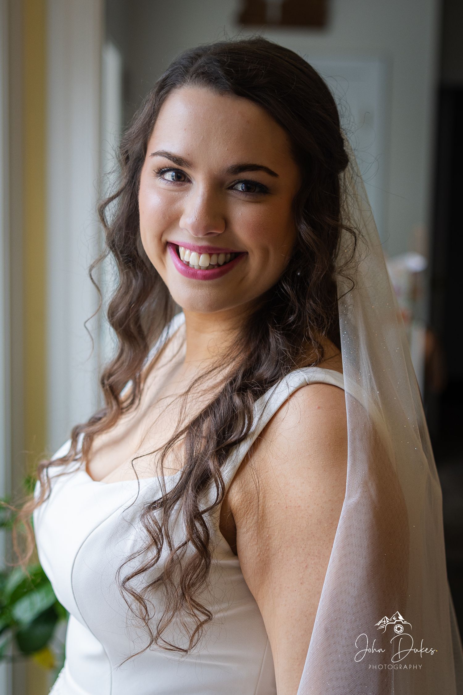 A bride in a white dress poses indoors with soft lighting and flowing curls.