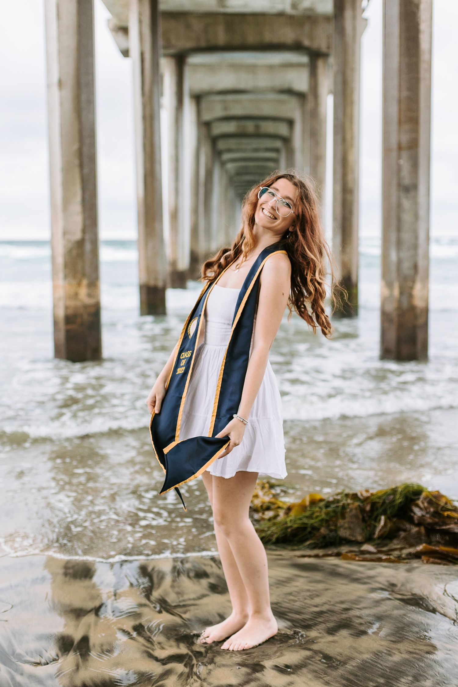 A woman wearing graduation attire stands and smiles underneath a pier with the ocean.