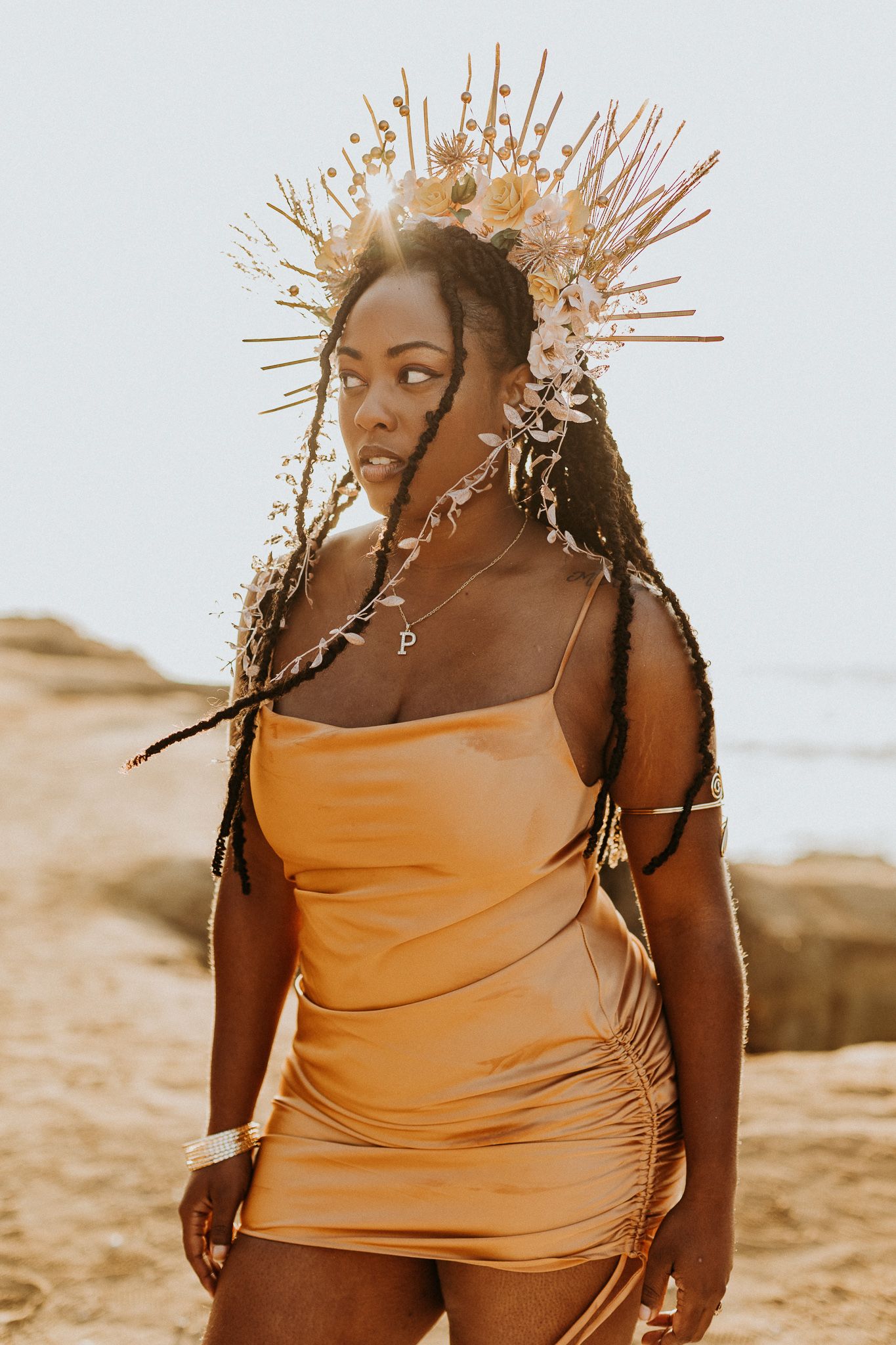 A woman wears a gold dress and dramatic flower crown during golden hour.