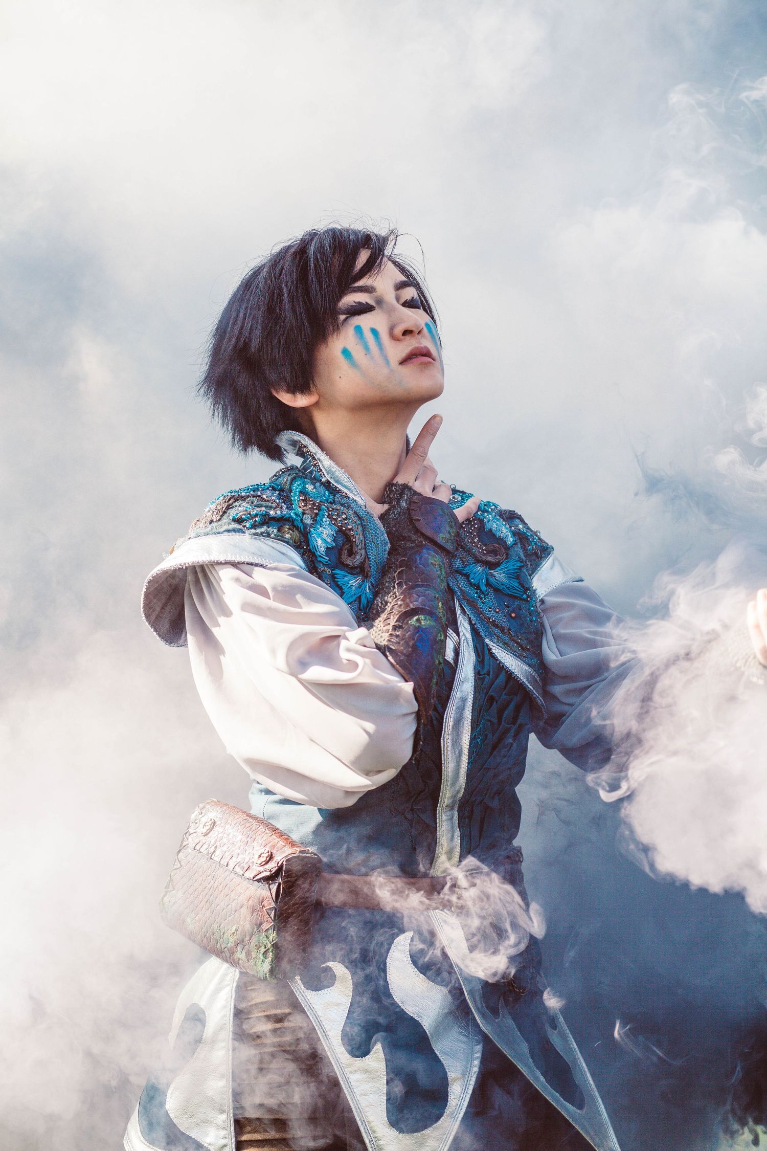 A woman wearing a blue cosplay outfit poses with eyes closed surrounded by smoke.