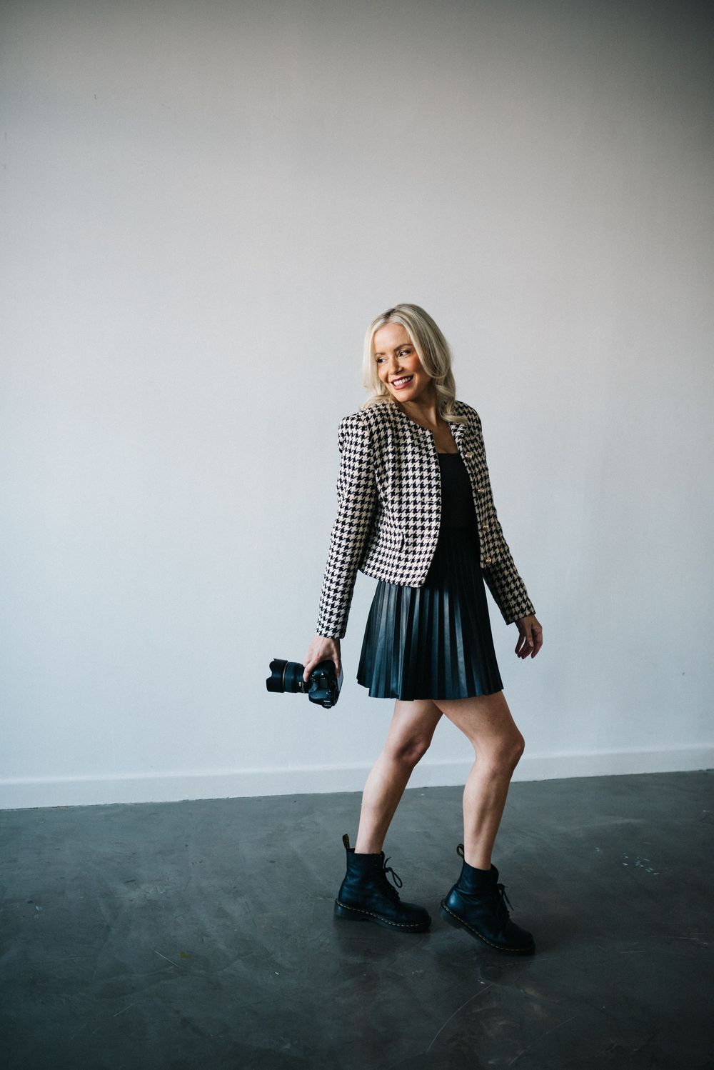 Smiling blonde woman in houndstooth blazer, black mini skirt, and boots holding a camera in a studio.