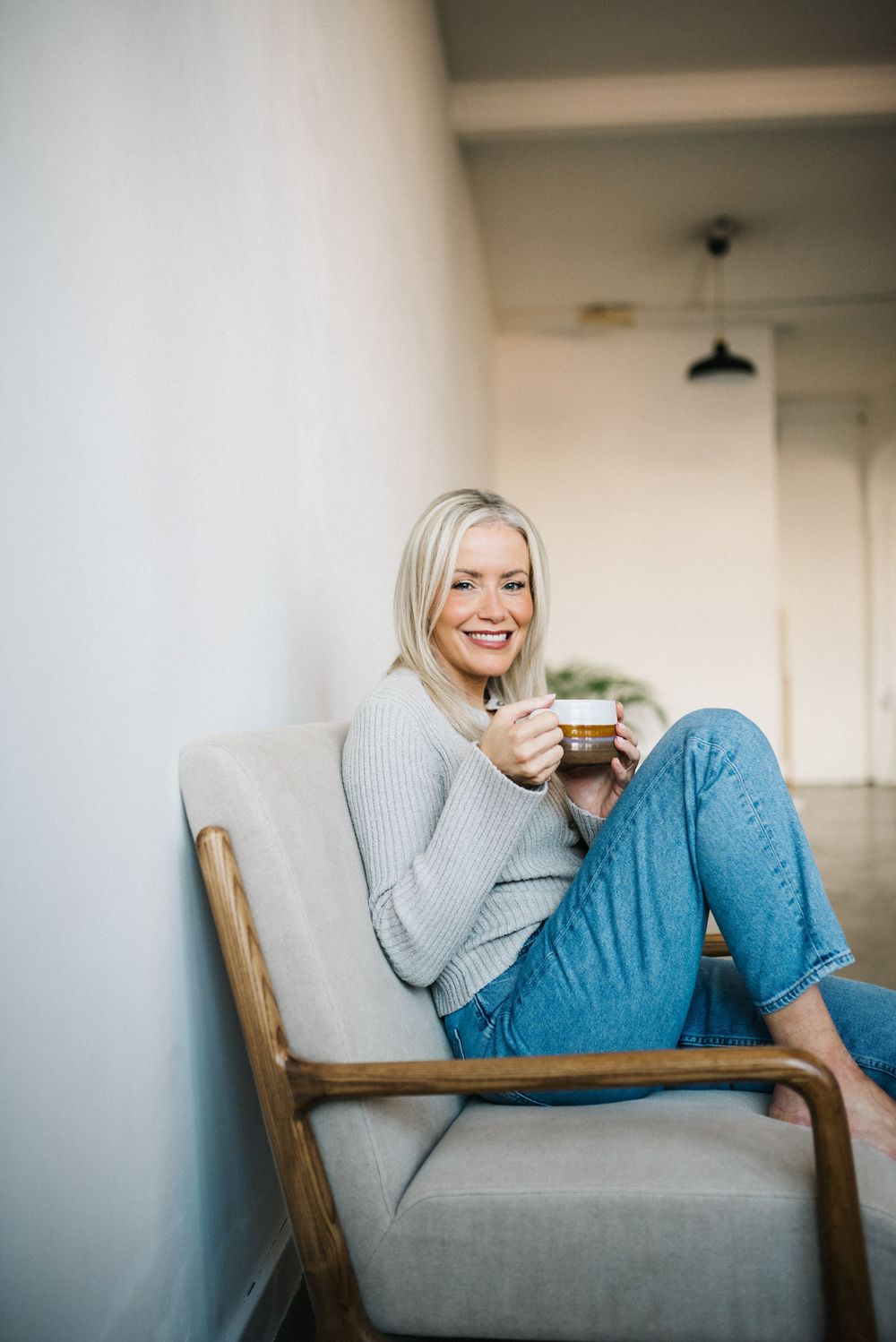 Smiling blonde woman relaxing in a chair holding a coffee mug, wearing jeans and a gray sweater.