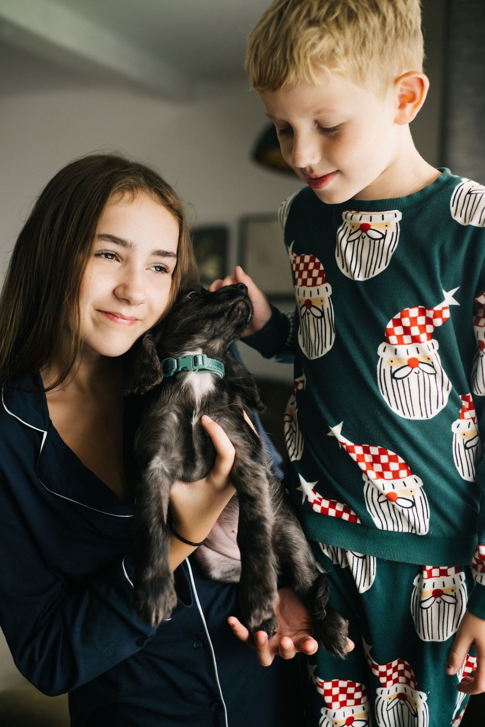 Two children in pajamas holding a small black puppy during Christmas morning celebrations at home.