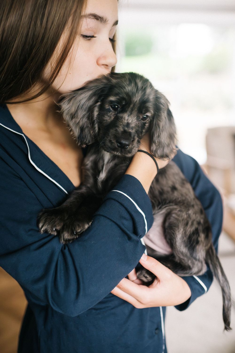 Young woman in navy pajamas kissing a fluffy black puppy she holds in her arms indoors.
