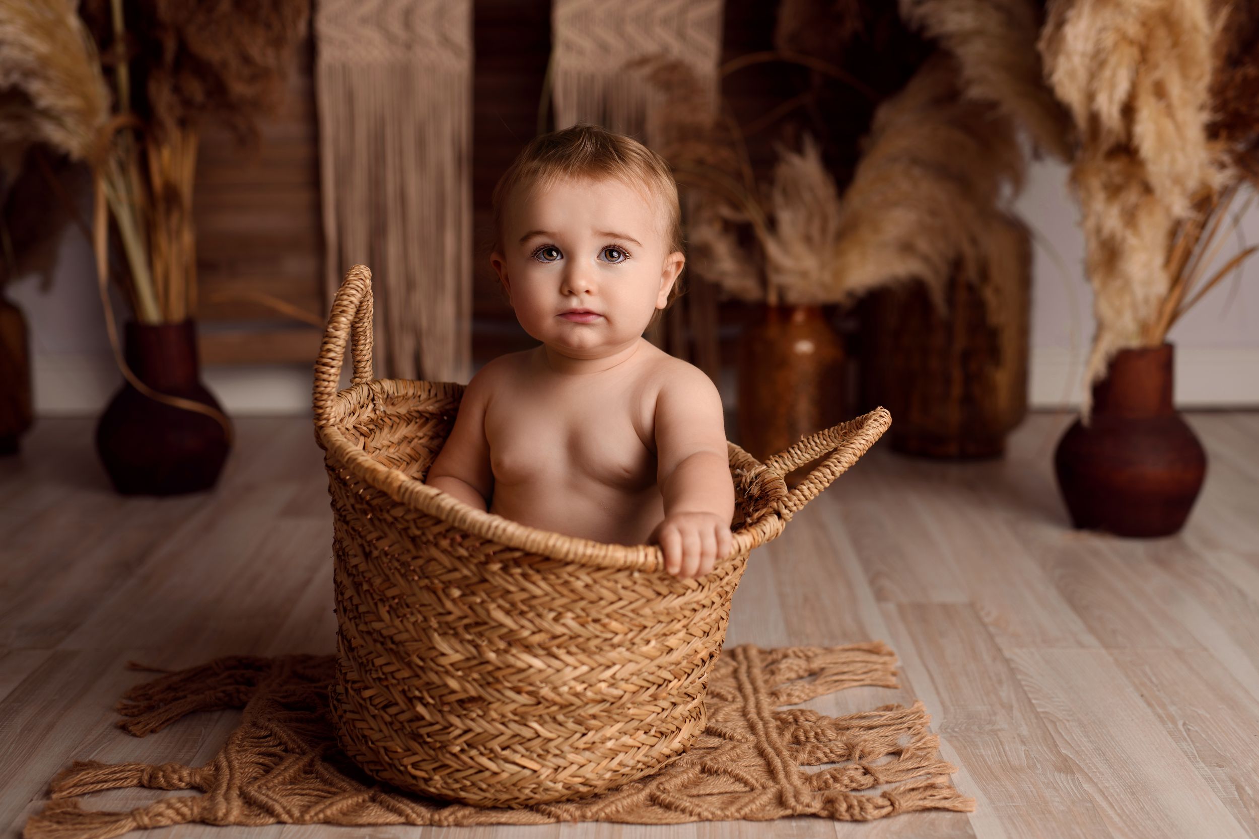 Cute baby sitting inside a large woven basket with rustic boho decor in background.