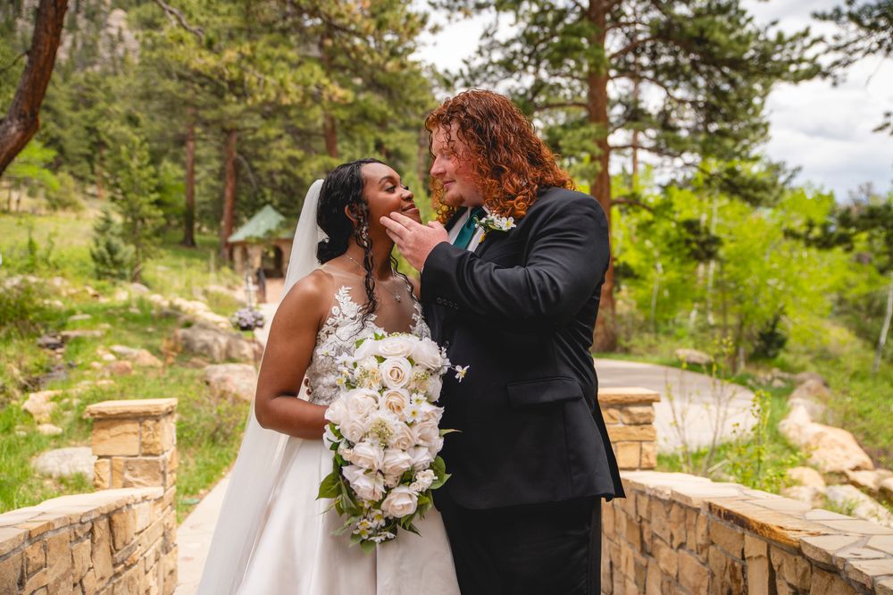 A couple shares an intimate moment on their wedding day in a beautiful outdoor mountain setting with stone walls and pine trees.