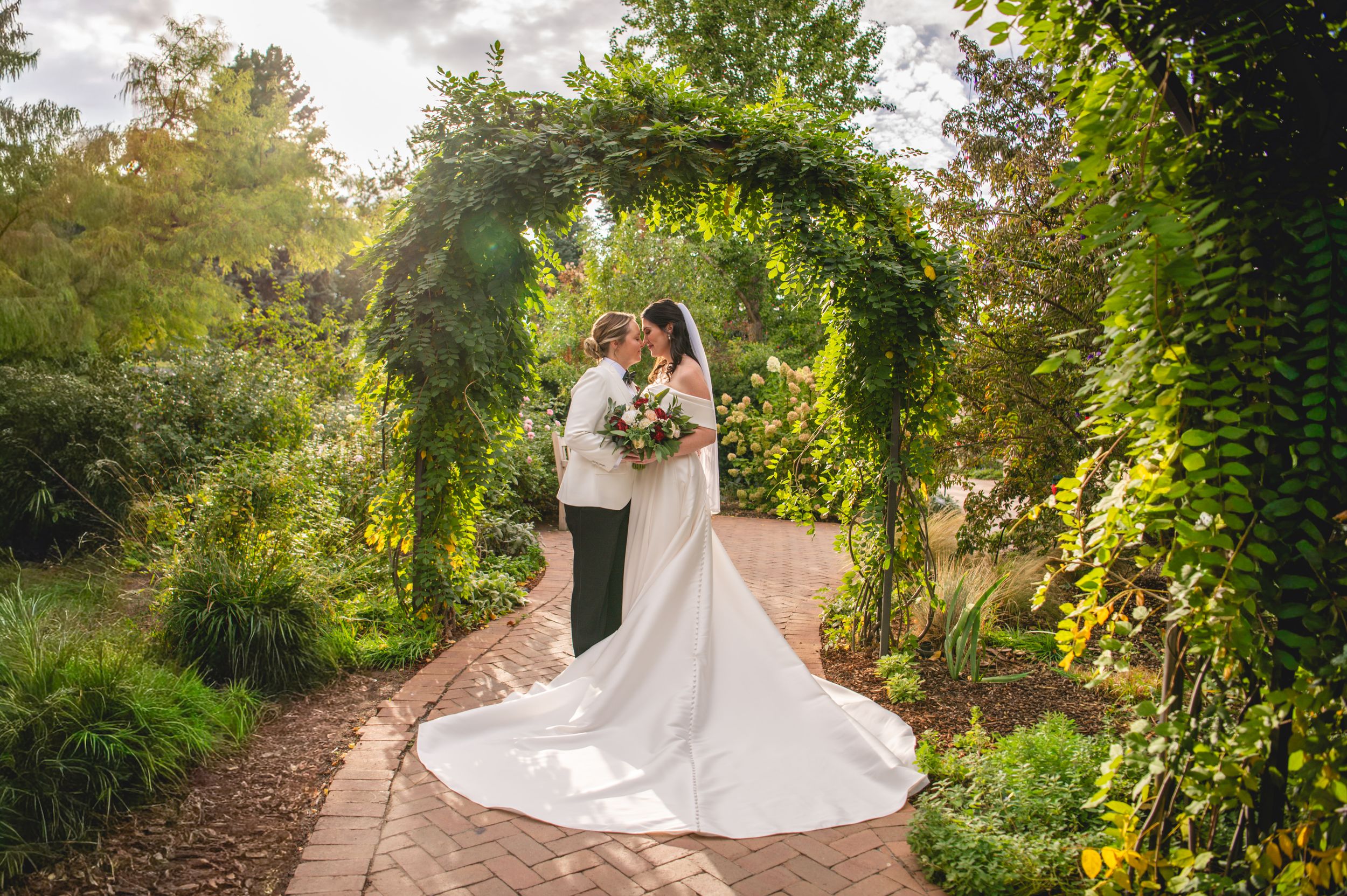 Bride in a long white gown and veil embraces her groom beneath a green ivy arch on a brick garden path at an outdoor wedding.