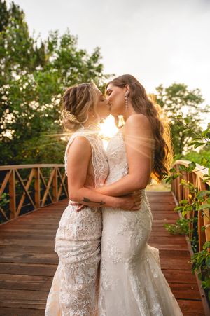 Two brides in white lace dresses share an intimate moment on a wooden deck at sunset.