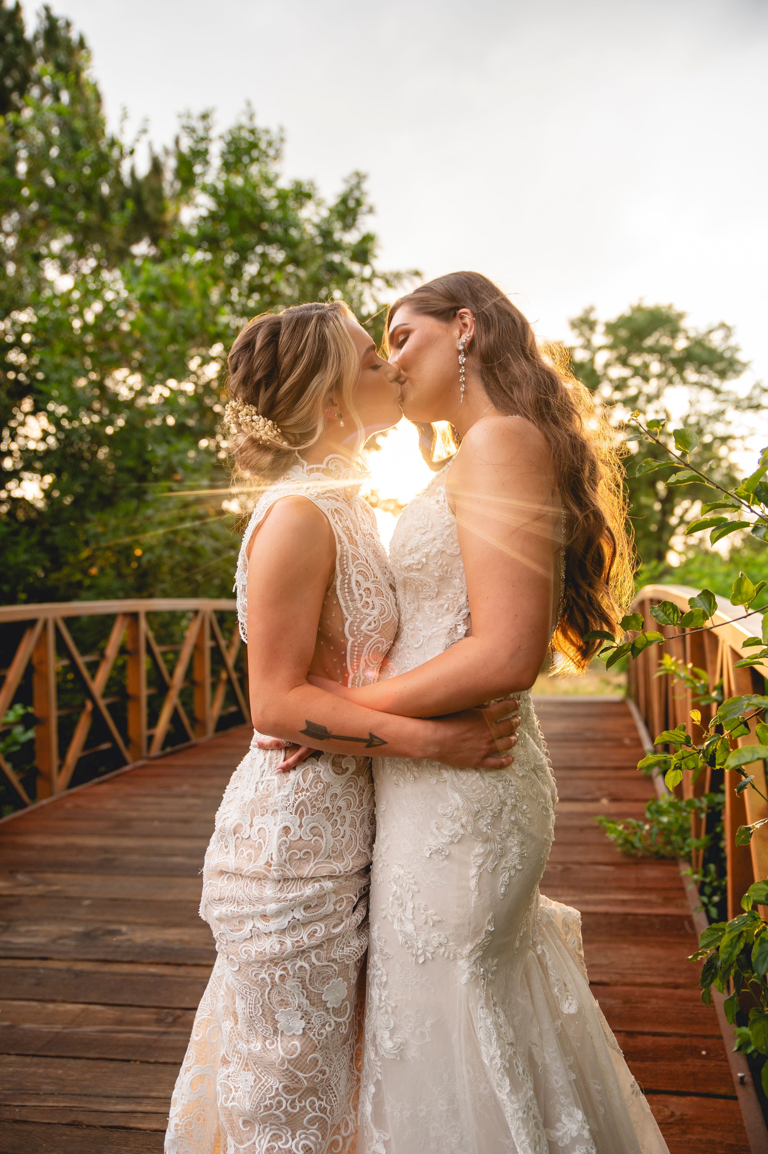 Two brides in white lace dresses share an intimate moment on a wooden deck at sunset.