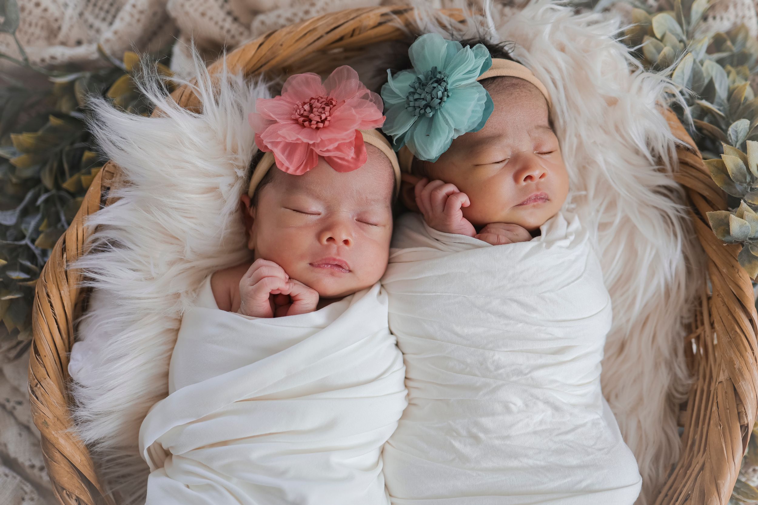 Two sleeping infants in white swaddles rest in a basket, adorned with pink and blue floral hair accessories.