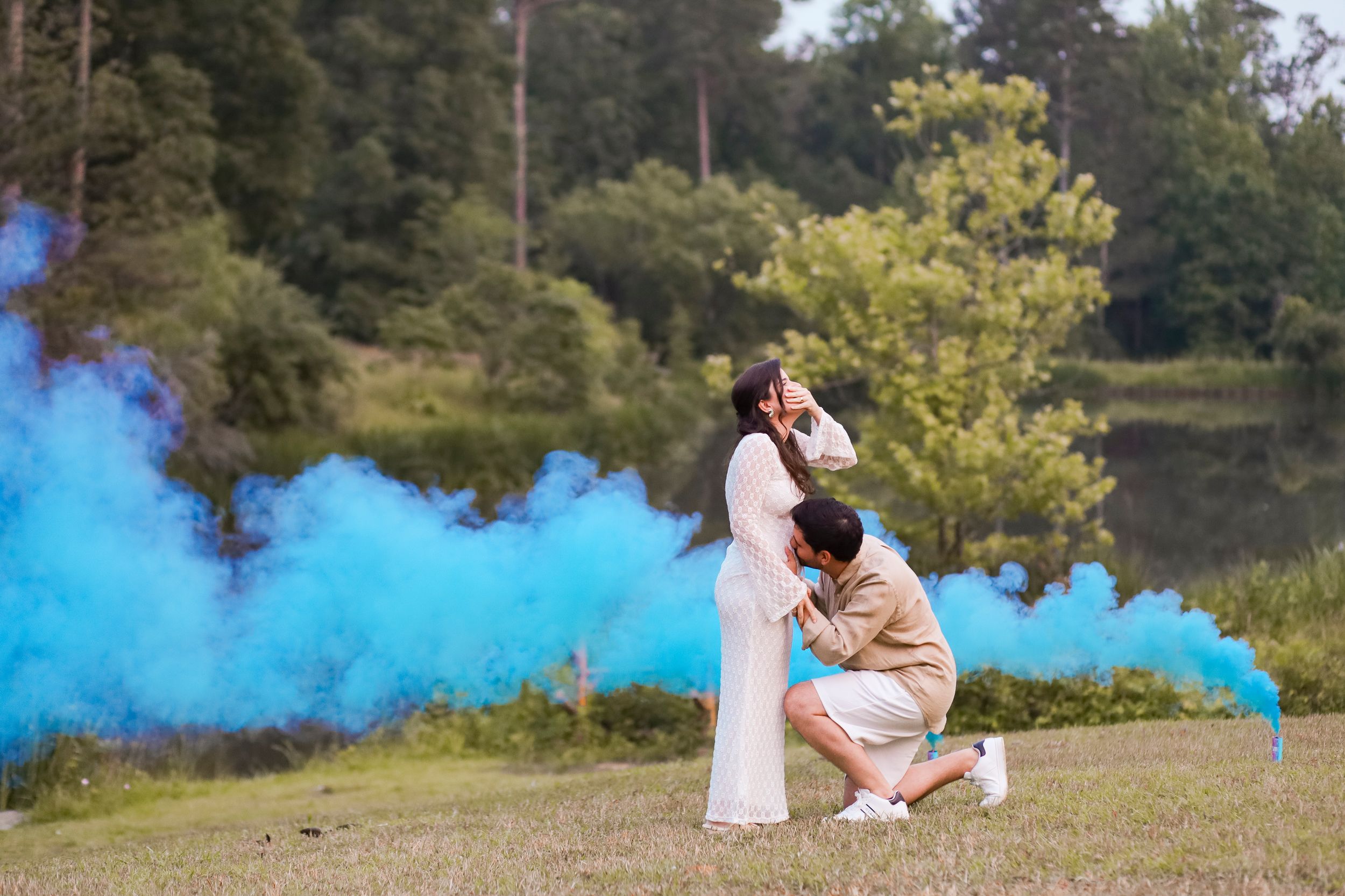 Couple in casual outfits sitting in grass with blue smoke bombs creating dreamy atmospheric backdrop at outdoor park setting.