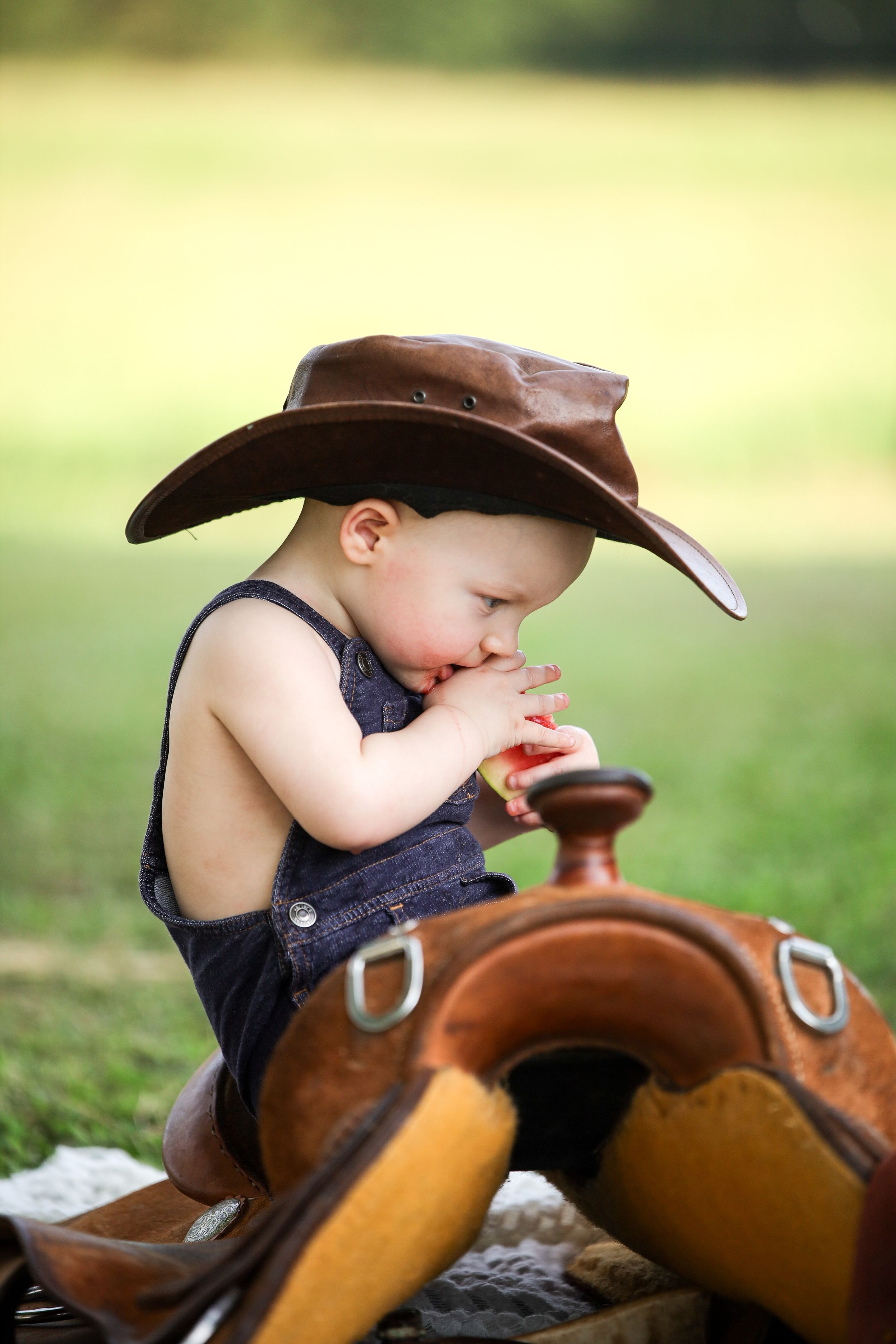 A small child in denim overalls and brown cowboy hat sits with a saddle on a sunny day outdoors.