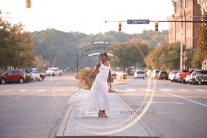 A person in a flowing white dress stands in the middle of a street at sunset while fabric billows in the breeze.