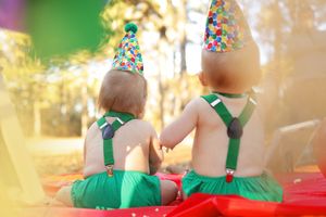 Two babies in green outfits and sparkly party hats sit together at a festive celebration with bokeh lights in background.
