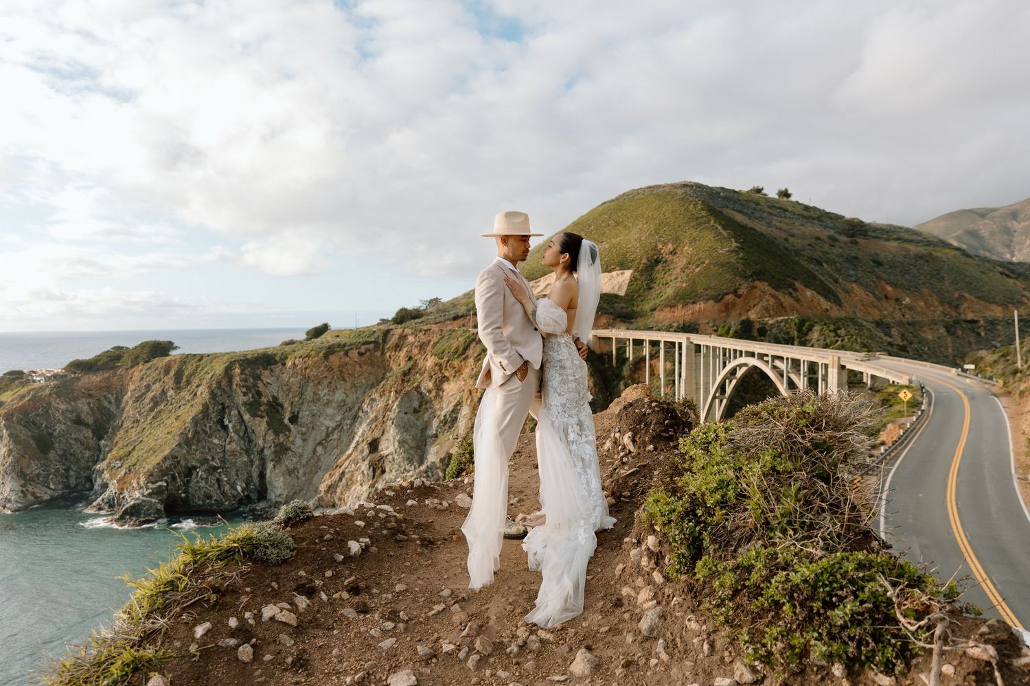Couple in white outfits standing near iconic Bixby Bridge in Big Sur, California.