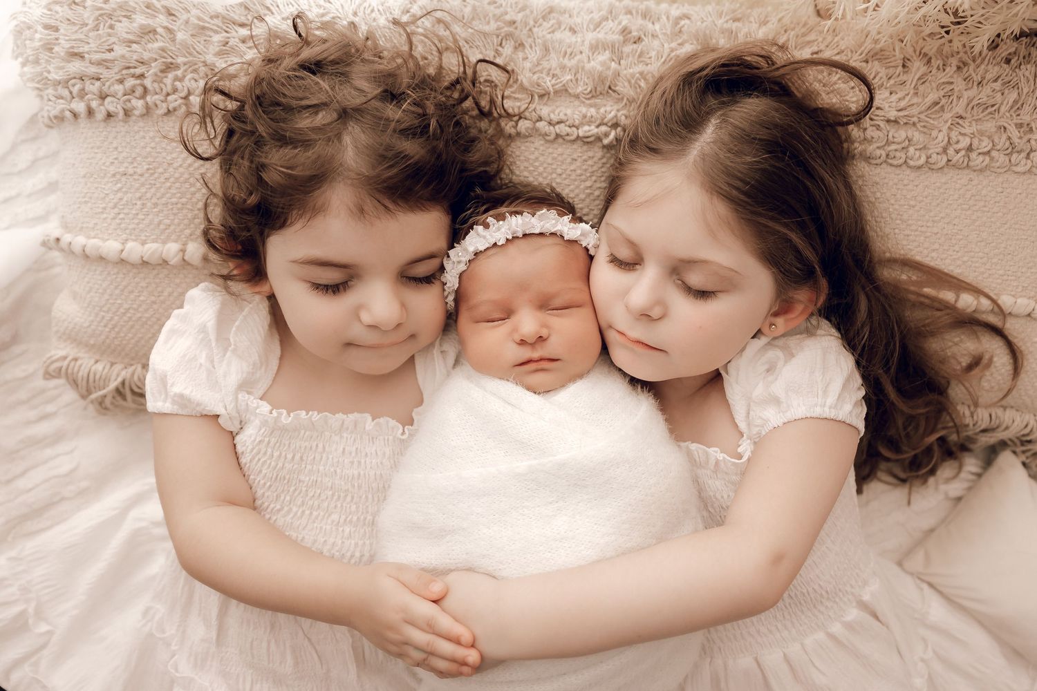 Two children in white dresses lying with swaddled newborn on cream textured bedding.