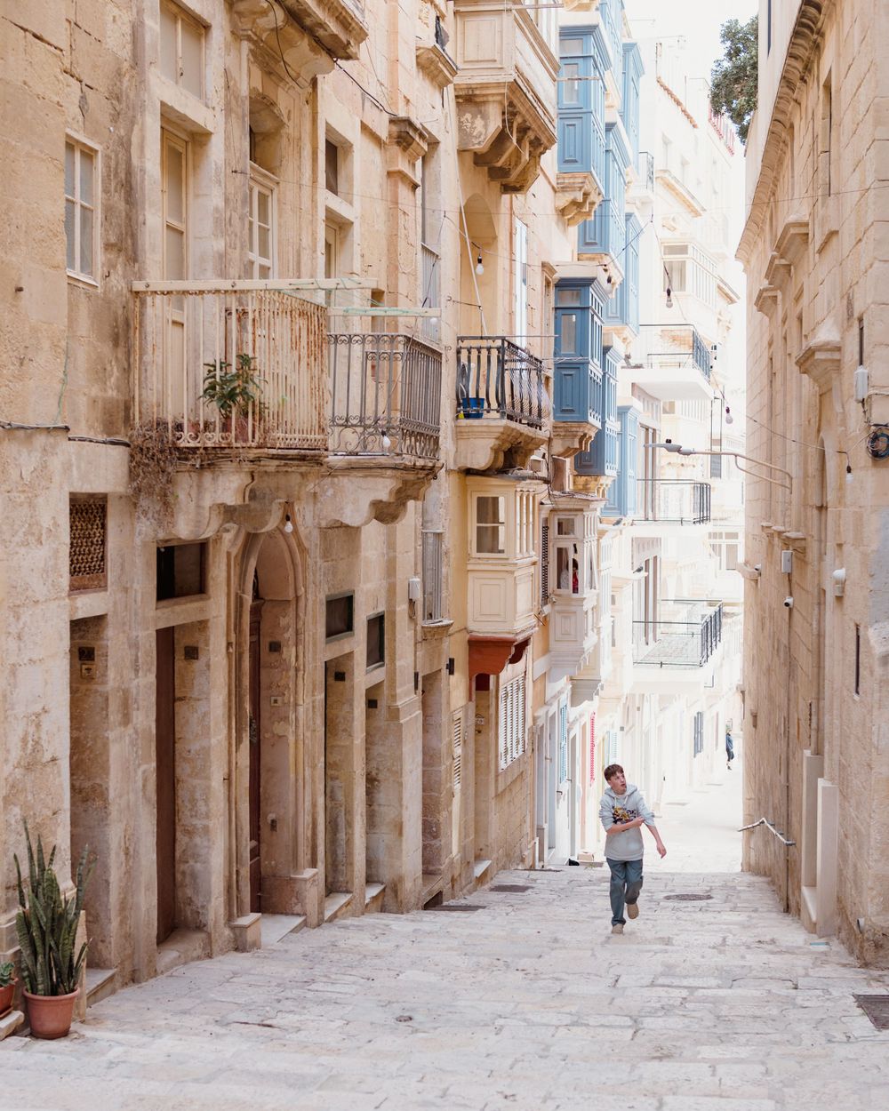 A person runs through a narrow stone alleyway lined with traditional balconies and architecture in Malta's historic streets.