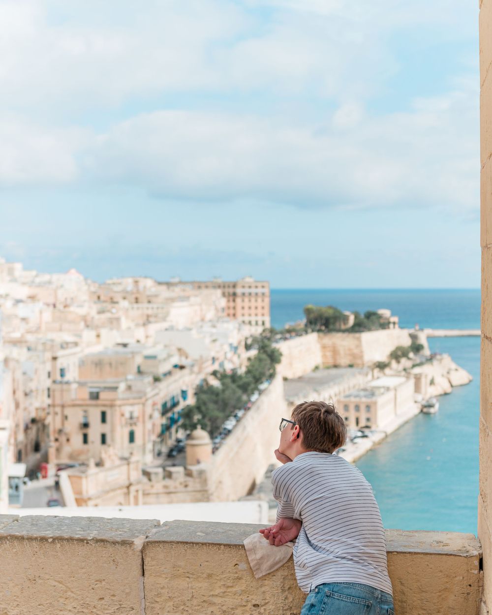 Person in striped shirt taking photos of a scenic coastal Mediterranean town with white buildings and blue waters.