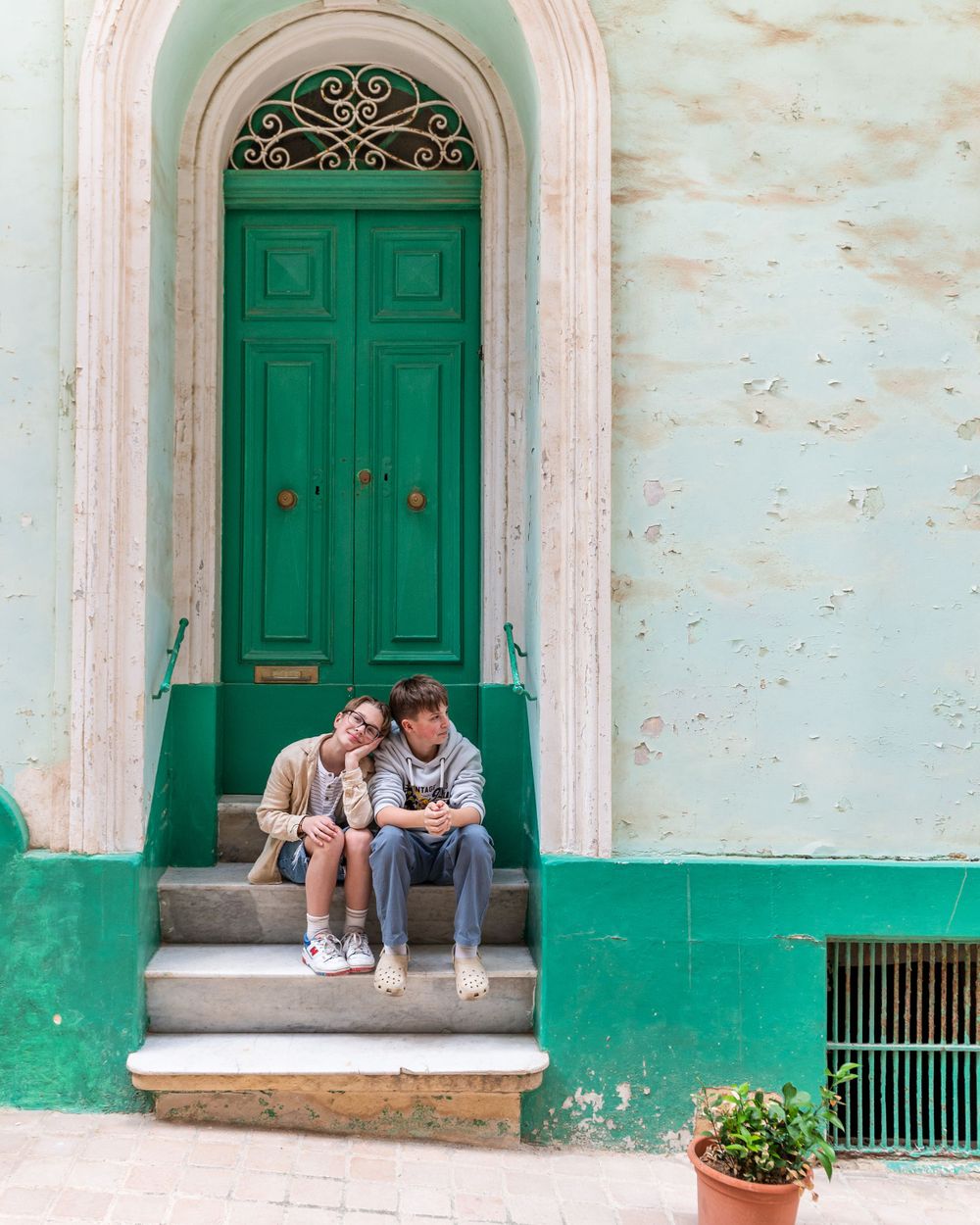 Two people sit together on the steps of a building with green doors and weathered white walls.