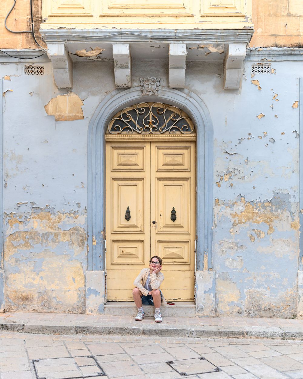 A person sits on a stone step against a weathered wall with peeling paint and yellow doors in an old building facade.