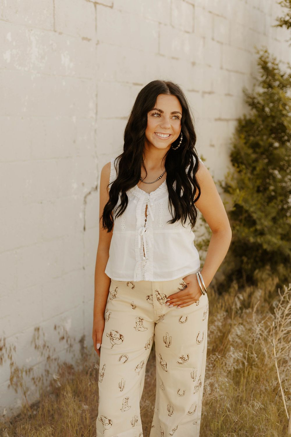 Western Fashionable outfit featuring a white sleeveless top and beige patterned pants against a white wall.