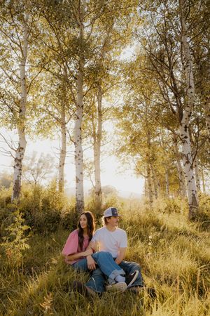 A western couple sits together in a golden-lit birch forest grove during sunset, surrounded by tall grass and dappled light.