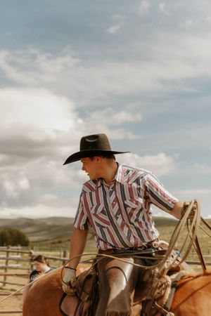 A cowboy in a striped shirt rides a horse across a ranch with mountains in the background. Family Branding