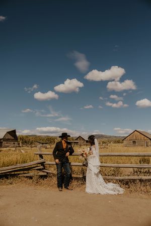 A western-themed wedding couple poses near rustic wooden structures under blue skies.
