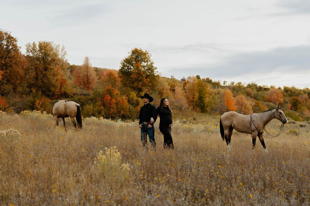 Couple standing together in autumn field with two horses grazing among golden grasses and colorful fall trees.