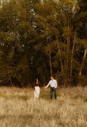 A western couple walking through tall grass at golden hour with trees in the background.