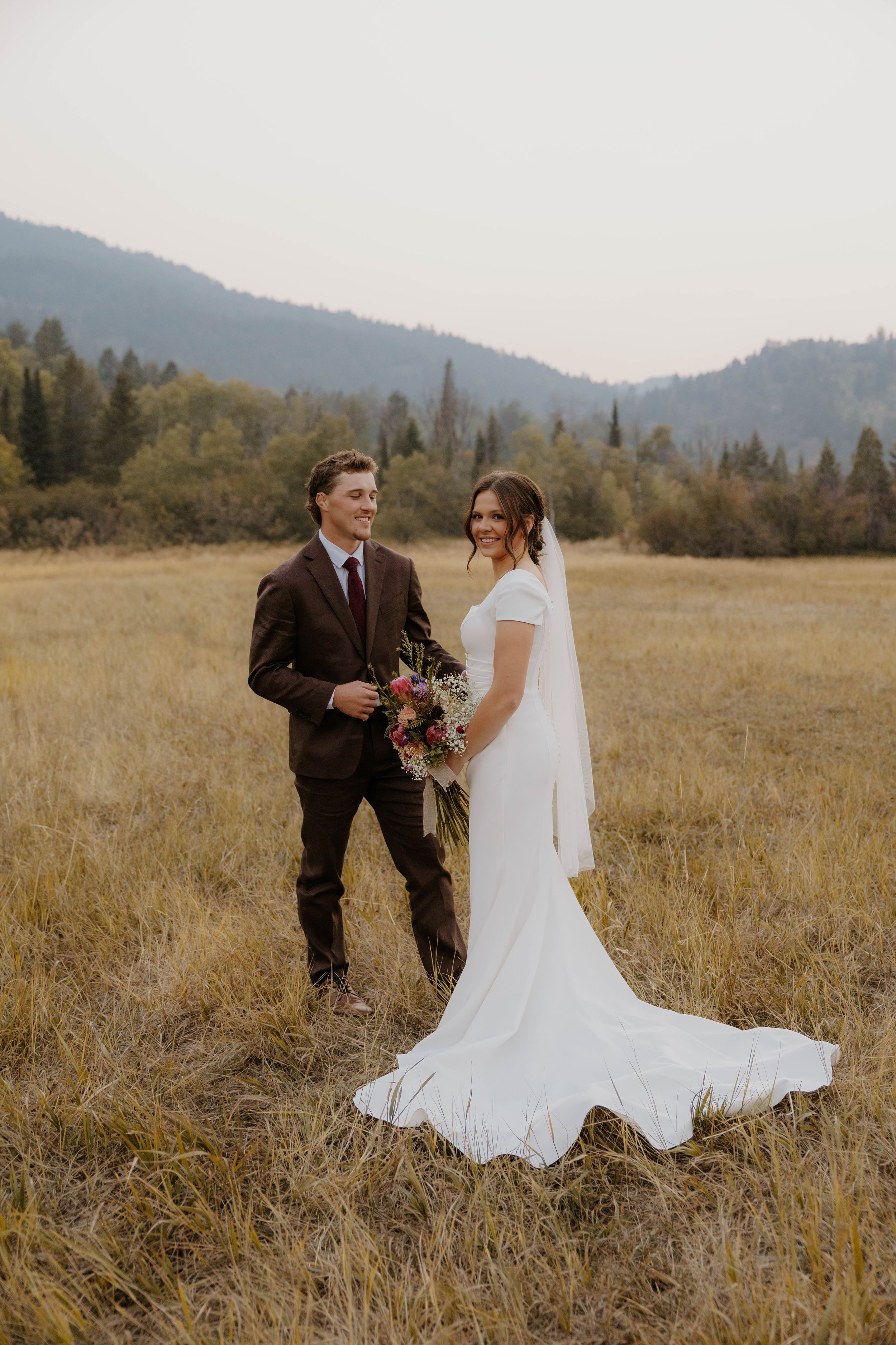 Bride in white gown with veil holds bouquet beside groom in brown suit in a golden meadow with mountain backdrop.