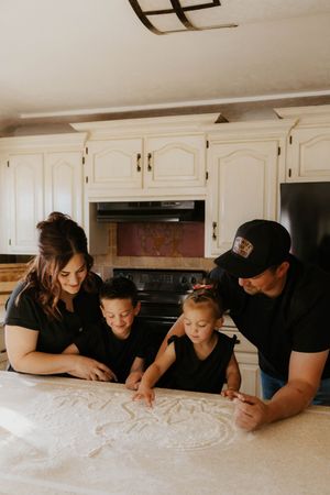 Family of four in matching black outfits gathered around a kitchen counter, drawing in flour together.